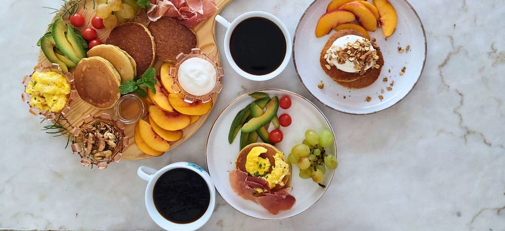 Chávenas de café, fruta, ovos mexidos e presunto , tomate cherry, frutos secos, e iogurte dispostos para um pequeno-almoço saudável