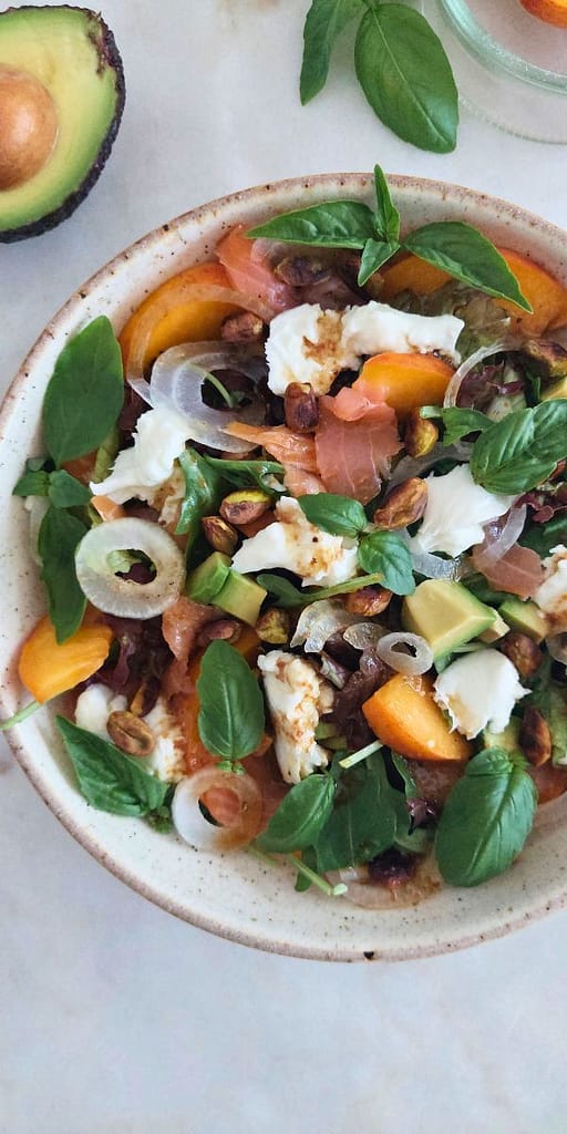Salada fresca de pêssego, salmão fumado e burrata com pistácios servida num prato grande, vista de cima