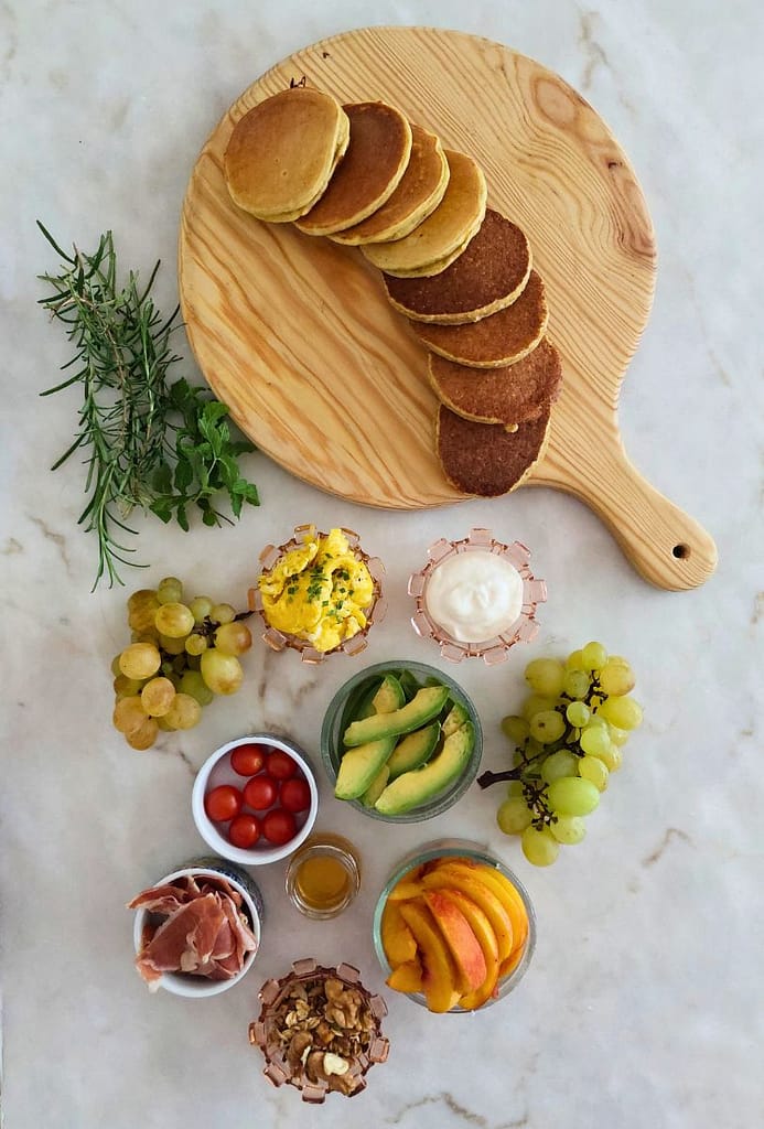 Panquecas, fruta, ovos mexidos e presunto, iogurte e mel ervas aromáticas e frutos secos disposto para montar Tábua de panquecas para brunch