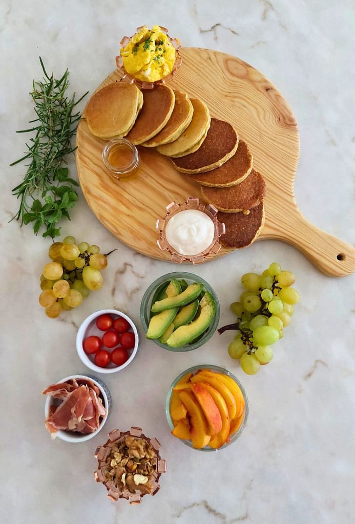 Panquecas, fruta, ovos mexidos e presunto, iogurte e mel disposto na bancada para montar uma tábua de panquecas para brunch