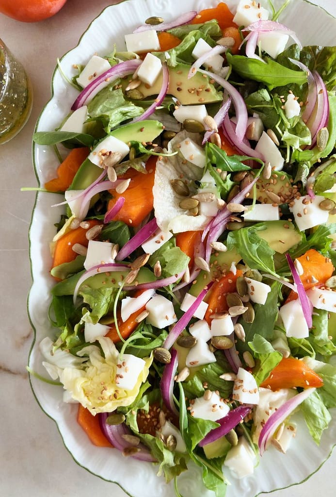 Salada colorida de diospiro tipo maçã com queijo fresco, abacate e sementes numa travessa polvilhada co sementes tostadas